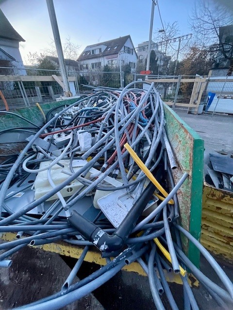 Im Vordergrund steht ein Container, der gefüllt ist mit verschiedenen Plastikrohren und Plastikteilen, die bei einem Gebäudeumbau gesammelt werden.